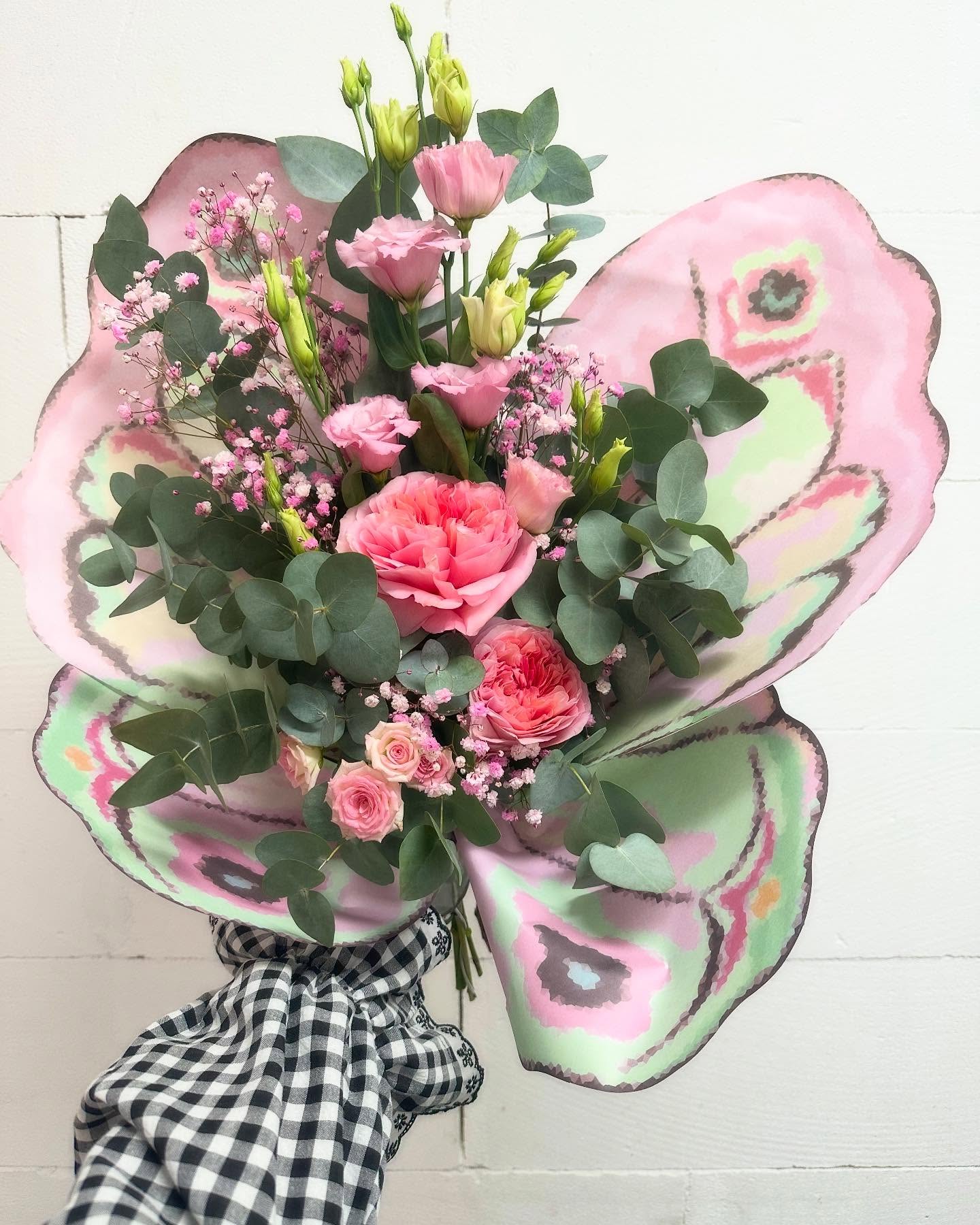 Bouquet de mariée papillon aux tons roses Bouquet de mariée en forme de papillon, composé de roses et lisianthus roses, gypsophile et eucalyptus, présenté dans un emballage artistique rose et vert en forme d’ailes. Une création florale originale et élégante pour un mariage unique.