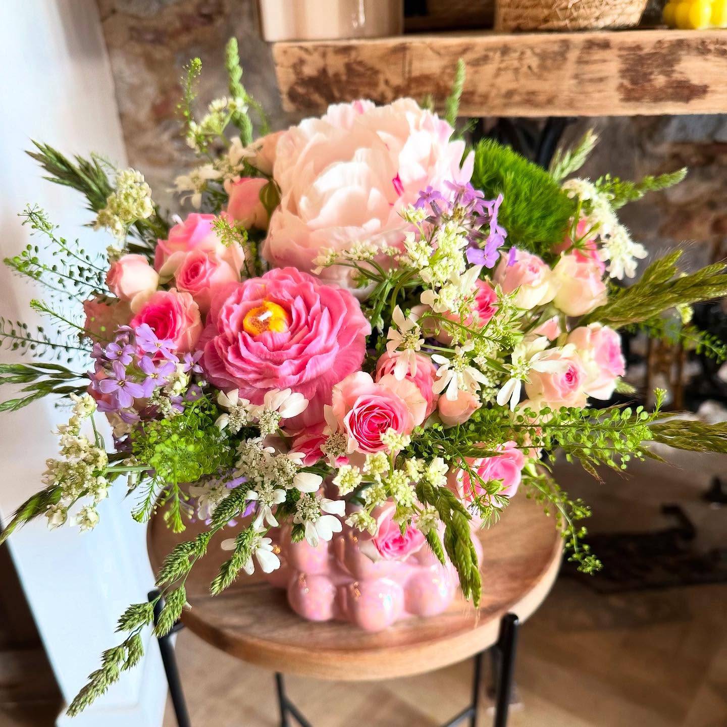 Bouquet de fleurs de saison aux tons roses et blancs Bouquet de fleurs de saison composé de pivoines, de roses, de fleurs champêtres blanches et violettes, associé à un feuillage vert léger. L’ensemble est présenté dans un vase rose en céramique, créant une composition fraîche et printanière.