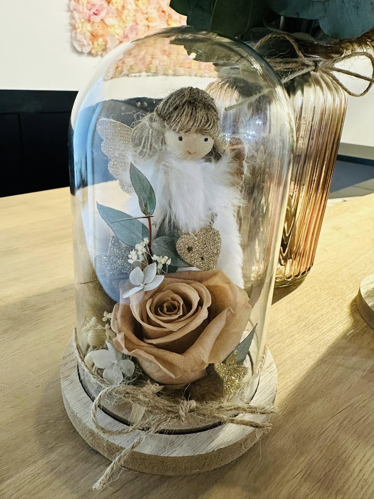 Cloche décorative avec rose séchée et petit ange en tissu Cloche en verre décorative contenant une rose séchée couleur pêche, des feuillages, des fleurs séchées blanches et une figurine d’ange en tissu avec des ailes scintillantes, posée sur une base en bois.